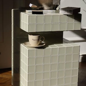 Op de afbeelding: Een witte betegelde bijzettafel met twee planken. De bovenste plank heeft een klein wit schaaltje met een bruin voorwerp erin, een klein zwart rechthoekig voorwerp en een witte doos met zwarte tekst. De onderste plank heeft een witte mok met het woord "Cappuccino" erop en een witte schotel.