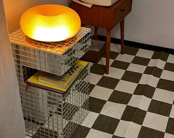 May include: A modern interior scene featuring a mirrored side table with a glowing orange donut-shaped lamp. A second mirrored table holds books. A wooden nightstand with a drawer and a box sits in the background. The floor has a black and white checkered pattern.