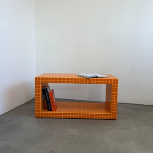 Puede incluir: Una mesa rectangular naranja con un patrón de cuadrícula y un estante abierto. Dos libros están dentro del estante y un libro está encima. La mesa está en una habitación con paredes blancas y suelo gris.