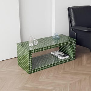 May include: A green tiled coffee table with a rectangular shape and a lower shelf. The table has a grid pattern and is paired with a black armchair and a wooden floor. A book and glassware are on the table.