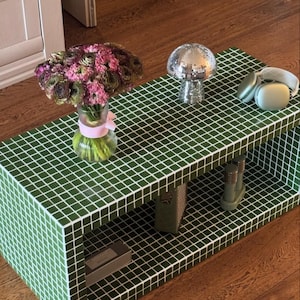 Puede incluir: Una mesa de café de azulejos verdes con dos estantes. La mesa tiene un jarrón de flores rosas y verdes, un hongo de bola de disco plateado y un par de auriculares azules claros.