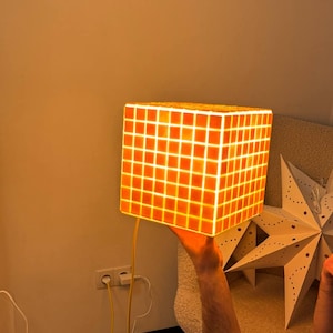 May include: A glowing orange cube lamp with a grid pattern. The lamp is held up by a person's hand. The lamp has a yellow power cord. The background is a neutral color.