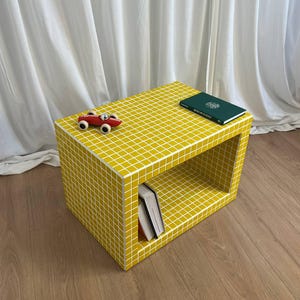 May include: A yellow, grid-patterned side table with a rectangular shape. A small red toy car sits on top, along with a green book. Two books are visible inside the open shelf. The table is on a wooden floor.