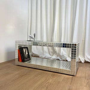 Puede incluir: Una mesa de centro rectangular espejada con un patrón de cuadrícula, que contiene dos libros y un libro abierto. La mesa está sobre un suelo de madera, con una cortina blanca al fondo.