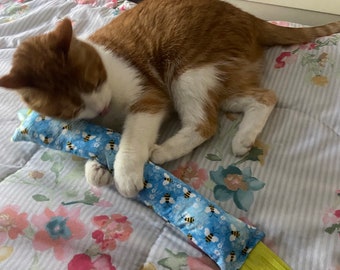 May include: An orange and white cat is lying on a floral patterned bedspread, playing with a blue cat toy. The toy is covered in a bee pattern and has a yellow fringe at the end.