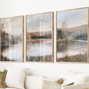 May include: A set of three vertical wall art prints in light wood frames, each depicting a serene landscape with a calm lake, soft-toned trees, and rolling hills in muted shades of brown, beige, and pale blue, arranged side by side on a wall.