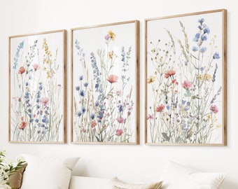 May include: Three framed watercolor paintings of a field of wildflowers in shades of blue, pink, and yellow. The paintings are in wooden frames and are hung on a white wall.