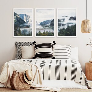 May include: Three framed landscape photographs of a mountain range with fog and trees. The photos are hung on a white wall above a bed with a white and black striped blanket and pillows.