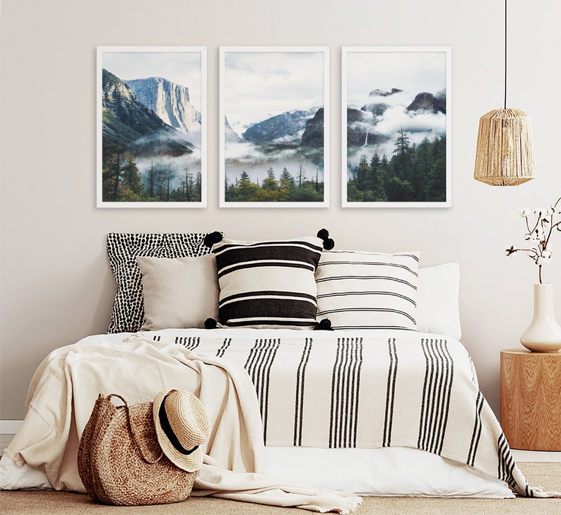 May include: Three framed landscape photographs of a mountain range with fog and trees. The photos are hung on a white wall above a bed with a white and black striped blanket and pillows.