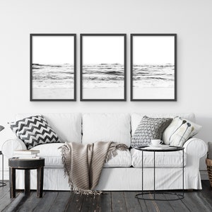 May include: Three black and white framed photographs of ocean waves crashing on a sandy beach. The photos are hung on a white wall above a white couch with pillows and a throw blanket.