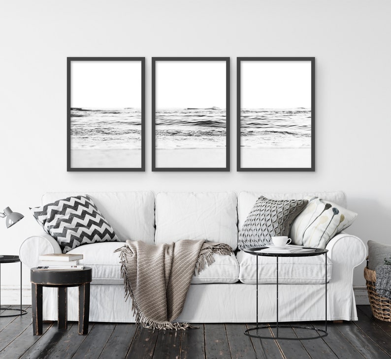 May include: Three black and white framed photographs of ocean waves crashing on a sandy beach. The photos are hung on a white wall above a white couch with pillows and a throw blanket.