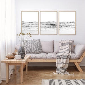 May include: Three black and white framed prints of ocean waves hanging on a white wall above a light gray couch with a striped throw blanket.