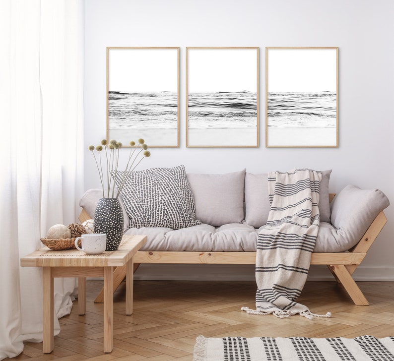 May include: Three black and white framed prints of ocean waves hanging on a white wall above a light gray couch with a striped throw blanket.