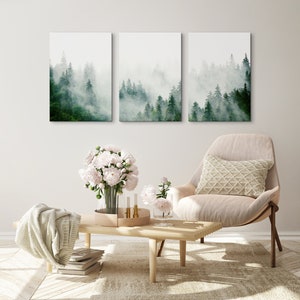 May include: Three framed prints of a misty forest scene with tall green trees. The prints are hanging on a white wall above a light brown wooden coffee table. A white armchair with a cream-colored pillow sits in front of the table. A bouquet of white peonies sits on the table.