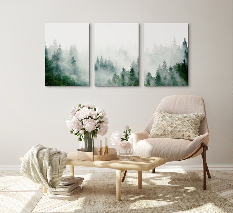 May include: Three framed prints of a misty forest scene with tall green trees. The prints are hanging on a white wall above a light brown wooden coffee table. A white armchair with a cream-colored pillow sits in front of the table. A bouquet of white peonies sits on the table.