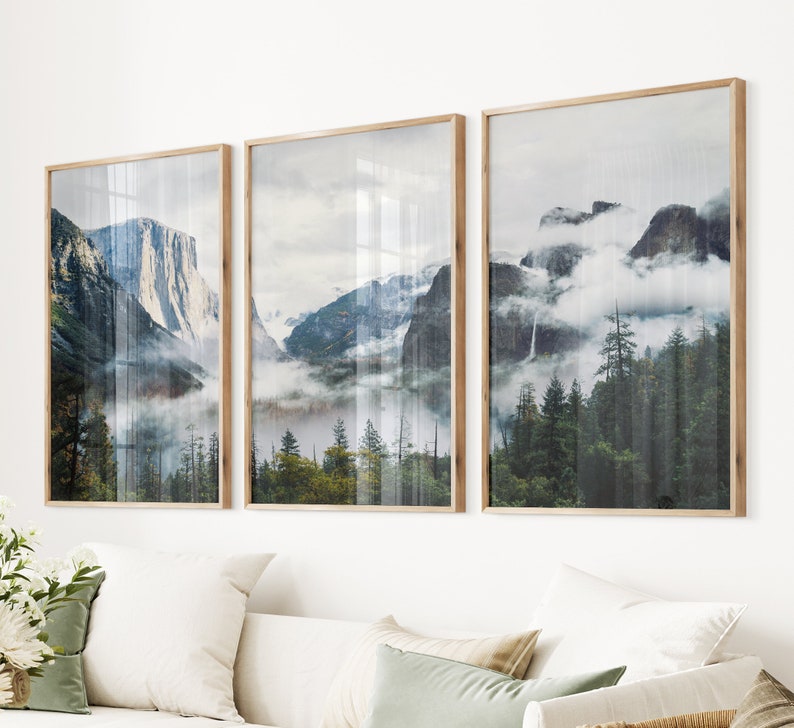 May include: Three framed landscape photographs of a mountain range with fog and trees. The photos are in wooden frames and are hung on a white wall.