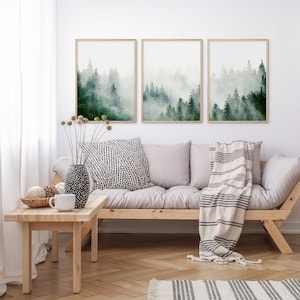 May include: Three framed prints of a misty forest scene with green trees. The prints are hanging on a white wall above a light gray couch with a white and black striped throw blanket.