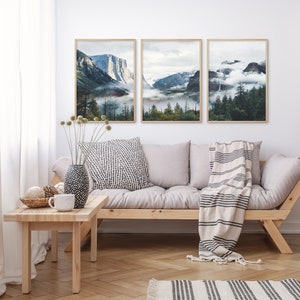 May include: Three framed landscape photographs of mountains and forests with fog and clouds. The photos are hanging on a white wall above a light grey couch with a white and black striped throw blanket.