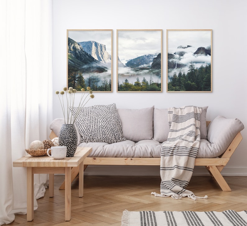 May include: Three framed landscape photographs of mountains and forests with fog and clouds. The photos are hanging on a white wall above a light grey couch with a white and black striped throw blanket.