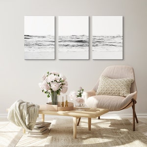 May include: Three black and white photographs of ocean waves crashing on a sandy beach. The photos are framed and hung on a white wall above a beige armchair with a white pillow. A bouquet of pink flowers sits on a wooden coffee table in front of the chair.