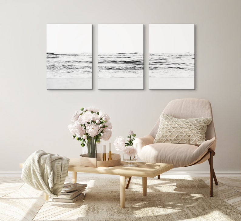 May include: Three black and white photographs of ocean waves crashing on a sandy beach. The photos are framed and hung on a white wall above a beige armchair with a white pillow. A bouquet of pink flowers sits on a wooden coffee table in front of the chair.