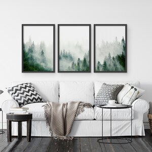 May include: Three framed prints of a misty forest scene with tall green trees. The prints are hung on a white wall above a white couch with pillows and a throw blanket.