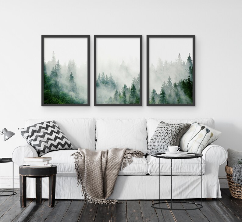May include: Three framed prints of a misty forest scene with tall green trees. The prints are hung on a white wall above a white couch with pillows and a throw blanket.
