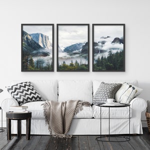 May include: Three framed landscape photographs of a mountain range with fog and trees. The photos are in black frames and are hung on a white wall above a white couch.
