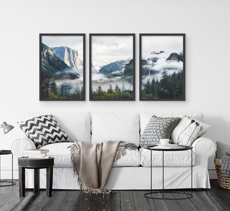May include: Three framed landscape photographs of a mountain range with fog and trees. The photos are in black frames and are hung on a white wall above a white couch.