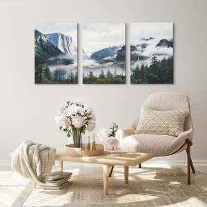 May include: Three framed landscape photographs of a mountain range with fog and trees. The photographs are hung on a white wall above a beige armchair with a white pillow.
