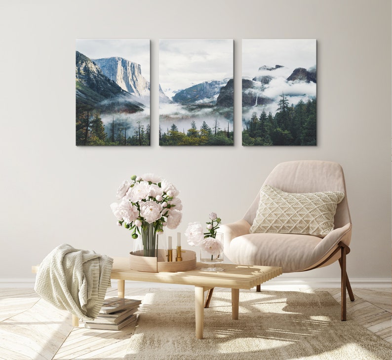 May include: Three framed landscape photographs of a mountain range with fog and trees. The photographs are hung on a white wall above a beige armchair with a white pillow.