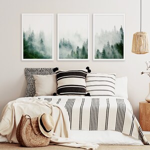 May include: Three framed prints of a misty forest scene with green trees and white fog. The prints are hanging on a white wall above a bed with a white and black striped blanket and pillows.