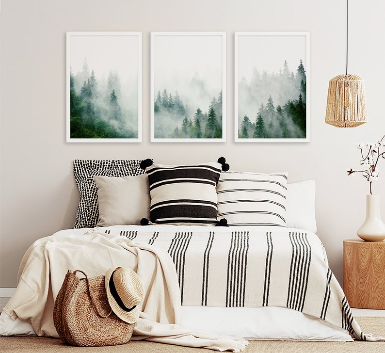 May include: Three framed prints of a misty forest scene with green trees and white fog. The prints are hanging on a white wall above a bed with a white and black striped blanket and pillows.