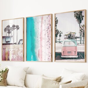 May include: Three framed prints with a beach theme. The left print shows a pink and blue beach scene with a surfboard. The middle print shows a blue ocean and a pink beach with people on it. The right print shows a pink and blue vintage van with surfboards on top of it.