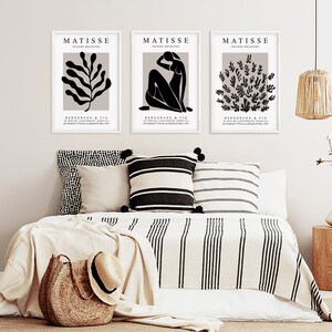 May include: Three black and white Matisse prints with the text "MATISSE PAPIERS DECOUPES" and "BERGGRUEN & CIE" framed in white and displayed on a white wall above a bed with a white and black striped blanket and pillows.