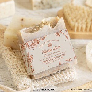 May include: A close-up of natural soap bars with a label that reads "Elysian Lore Charcoal Detox Soap". The soap is on a woven net, with a wooden brush, a loofah sponge, and other soap bars in the background. The soap is off-white with flecks.