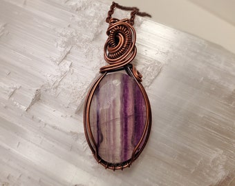 May include: A purple and white fluorite gemstone pendant wrapped in copper wire. The pendant is hanging from a copper chain.