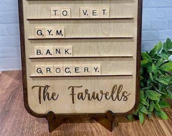 Könnte beinhalten: Ein Schild aus Holz mit braunem Rahmen und hellem Holzgrund. Das Schild enthält eine Liste von Aufgaben, die mit Scrabble-Steinen geschrieben sind. Die Aufgaben umfassen "Hund zum Tierarzt bringen", "Fitnessstudio", "Bank" und "Lebensmittelgeschäft". Das Schild trägt außerdem den Text "The Farwells" in goldenen Lettern am unteren Rand.