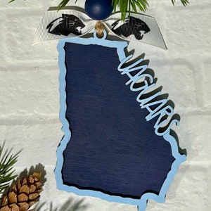 Puede incluir: Adorno de madera azul y blanco con forma del estado de Georgia con la palabra "Jaguars" escrita en él. El adorno está decorado con una cinta blanca con un logotipo de pantera negra.