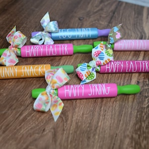 May include: Six wooden rolling pins decorated with colorful ribbons and painted with Easter-themed phrases, including "Happy Easter", "Bunny Snacks", and "Egg Hunt".