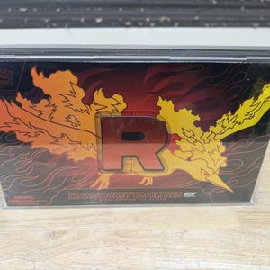 May include: A sealed Pokémon Ultra-Premium Collection box featuring a fiery Moltres design. The box has a large red "R" and the text "Team Rocket's Moltres EX". The background is a gradient of red and orange.