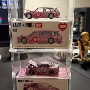 May include: Three pink die-cast model cars in a clear display case. The cars are a Nissan Skyline GT-R, a Nissan Skyline Wagon, and a Nissan Skyline Wagon. The cars are all in 1/28 scale and feature detailed paint and chrome accents. The display case is made of clear acrylic and has a black base.