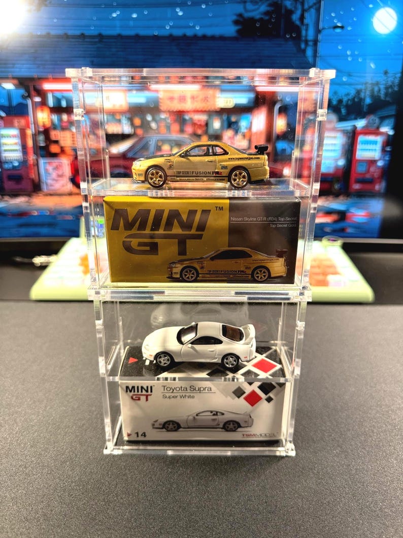 May include: Three miniature toy cars displayed in a clear acrylic case. The top car is a yellow Nissan Skyline GT-R with the words "IPDEEFUSION" printed on the side. The middle car is a white Nissan Skyline GT-R with the words "FUSIOMP" printed on the side. The bottom car is a white Toyota Supra with the words "Toyota Supra Super White" printed on the box. The box also has the words "MINI GT" printed on it.