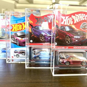 May include: Several Hot Wheels die-cast model cars in clear acrylic display cases. The cars are various colors, including purple, red, and blue. The packaging features the Hot Wheels logo and car model names.