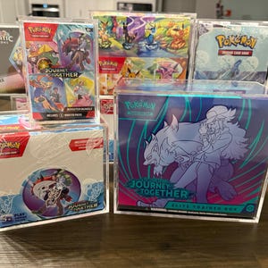 May include: A collection of Pokemon trading card game products, including a Journey to the Stars Elite Trainer Box, a Journey to the Stars booster bundle, and a Prismatic Evolutions surprise box. The boxes are clear plastic and feature artwork of Pokemon characters.