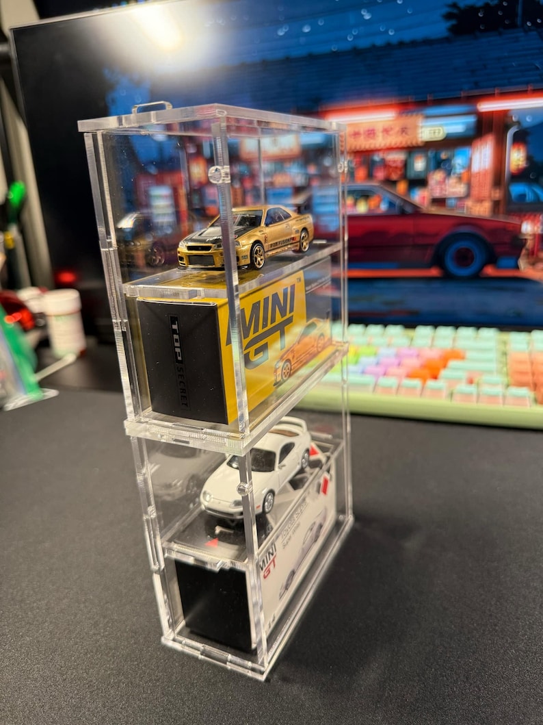 May include: A clear acrylic display case with three shelves, showcasing two miniature toy cars. The top shelf features a gold car with the text "MINI GT" on the box. The middle shelf features a white car with the text "TOP SECRET" on the box. The bottom shelf features a black box with the text "MINI GT" on it.