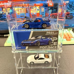 May include: A clear acrylic display case holding three miniature toy cars. The top shelf features a blue sports car with its hood open. The middle shelf has a box with a blue car image and text. The bottom shelf displays a white sports car.