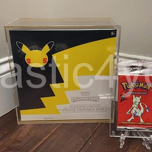 May include: A clear acrylic display case holding a Pokemon Celebrations Elite Trainer Box with a yellow and black design. A red Pokemon card pack is displayed on a clear stand. The box features a Pikachu graphic.
