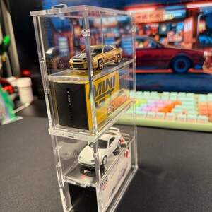 May include: A clear acrylic display case with three shelves, showcasing two miniature toy cars. The top shelf features a gold car with the text "MINI GT" on the box. The middle shelf features a white car with the text "TOP SECRET" on the box. The bottom shelf features a black box with the text "MINI GT" on it.