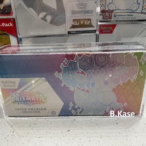 May include: A clear acrylic display case with a Pokemon card collection inside. The case features a holographic image of Eevee with a rainbow background. The text on the box reads "Sword & Shield Prismatic Evolutions Super-Premium Collection".